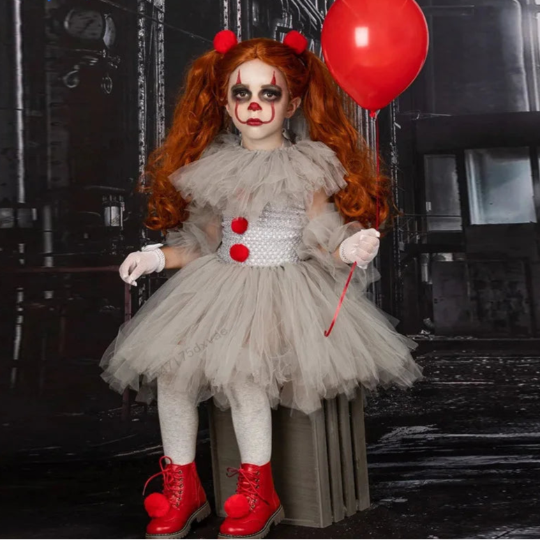 Person in a clown costume with red hair and makeup, holding a red balloon, against a dark background.