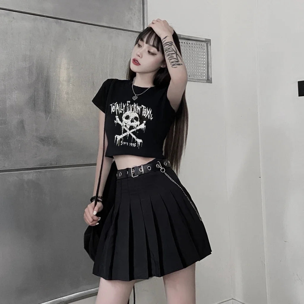 Person wearing a black t-shirt with a skull design and a black pleated skirt against a gray wall.